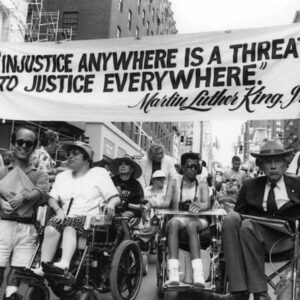  Photo of 1970s ADAPT demonstration where there is a row of people using wheelchairs on a city the street holding up a banner with the MKL quote “Injustice anywhere is a threat to justice everywhere.”